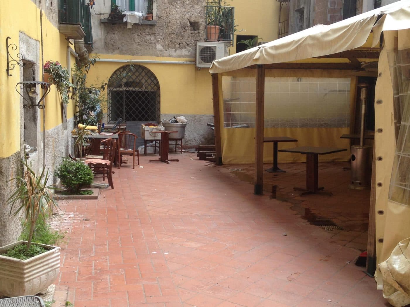 Il cortile prima dei lavori: muri gialli, pavimento di cotto e un gazebo chiuso dai teli