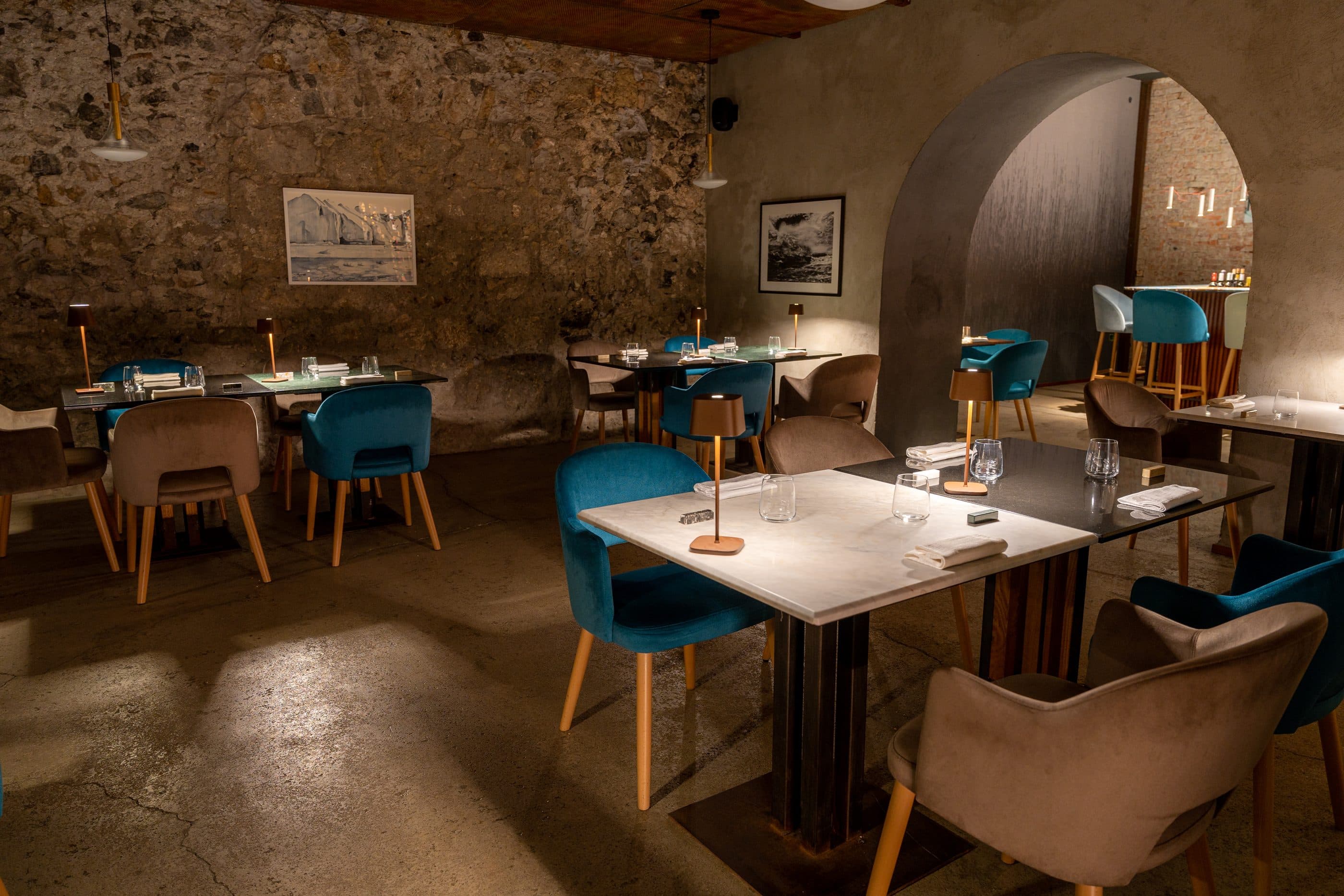 La sala interna con il muro di pietra a vista, i tavoli in marmo e le poltroncine di velluto petrolio