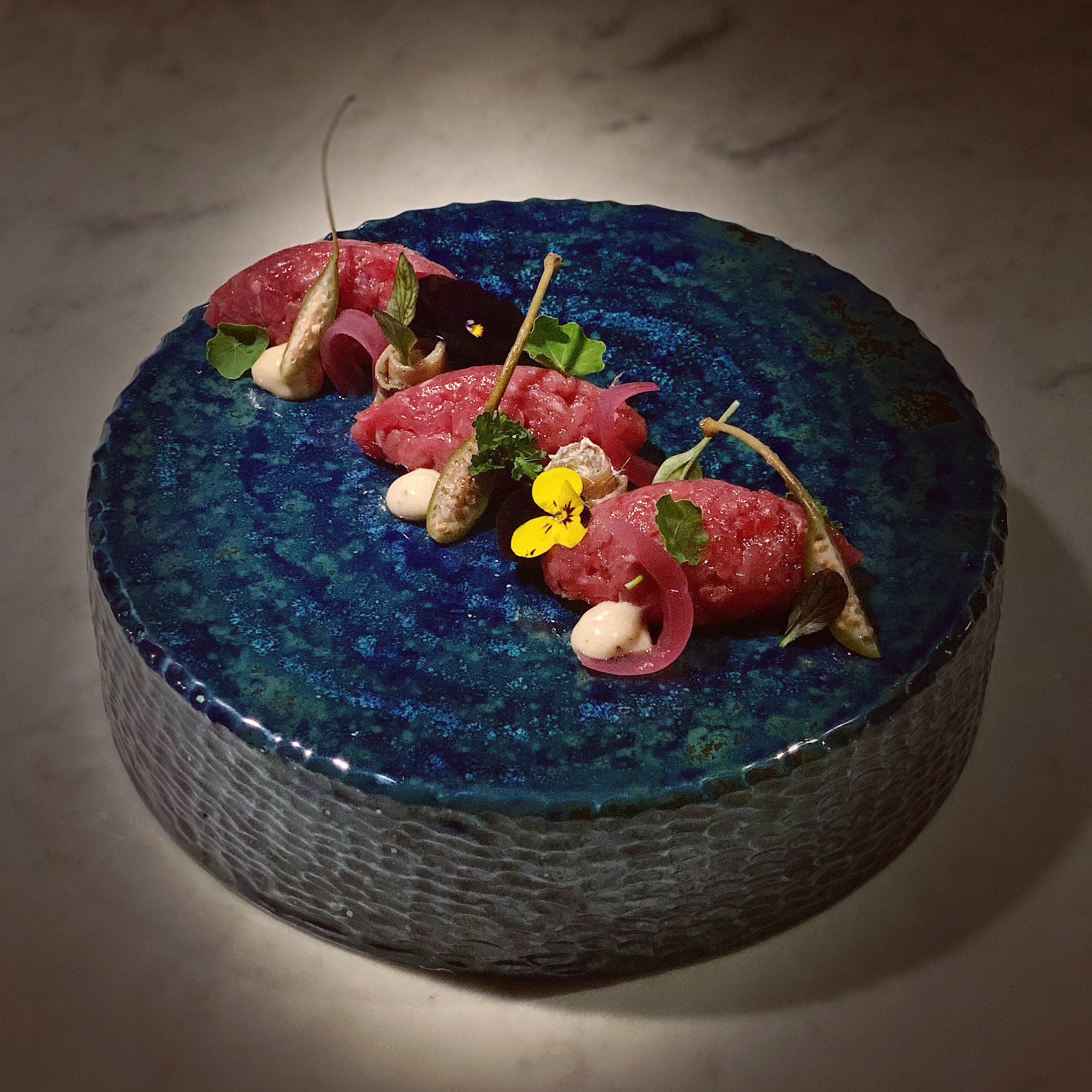 Tartare di Fassona servita su un piatto di ceramica blu smaltata, con fiori eduli e gocce di salsa